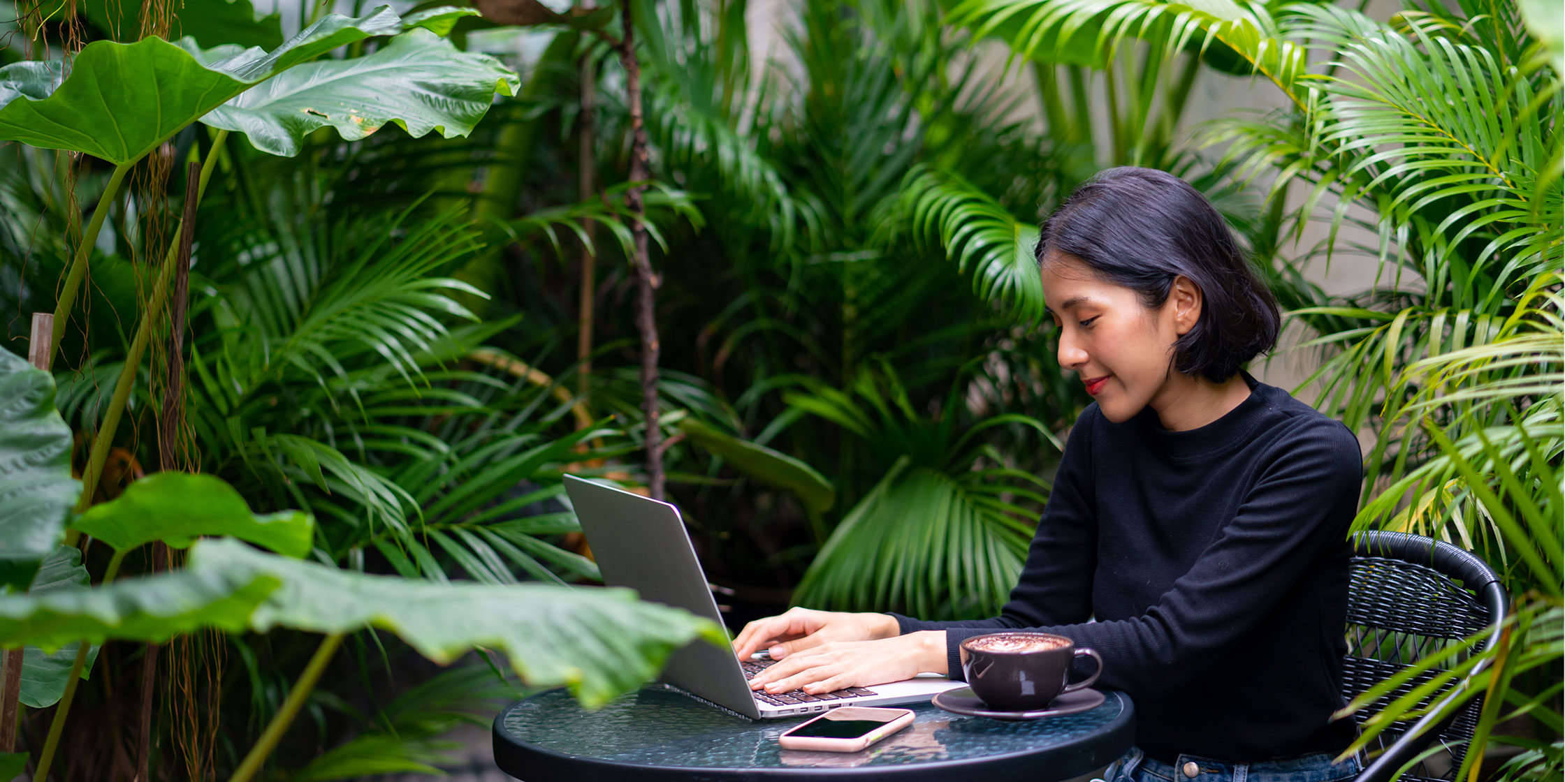 A person sitting at a table surrounded by green plants uses their laptop to check their credit limit.