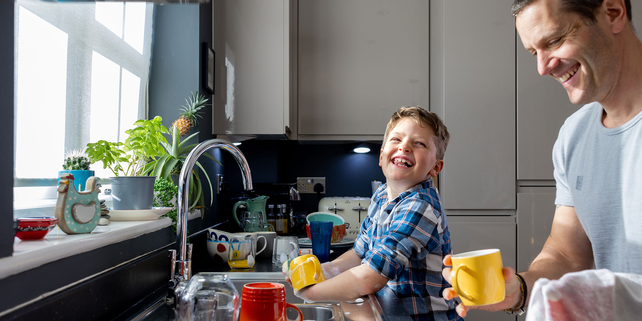 A child and an adult laugh together while cleaning dishes at the kitchen sink.