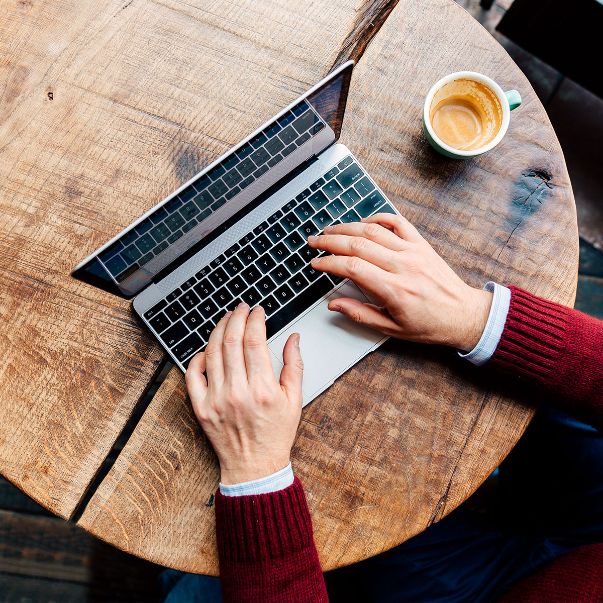 An overhead photo of a person’s hands typing on a laptop. There is a cup of coffee beside them.