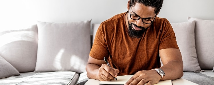 A man wearing glasses and a watch sits in the sun on his couch and writes.