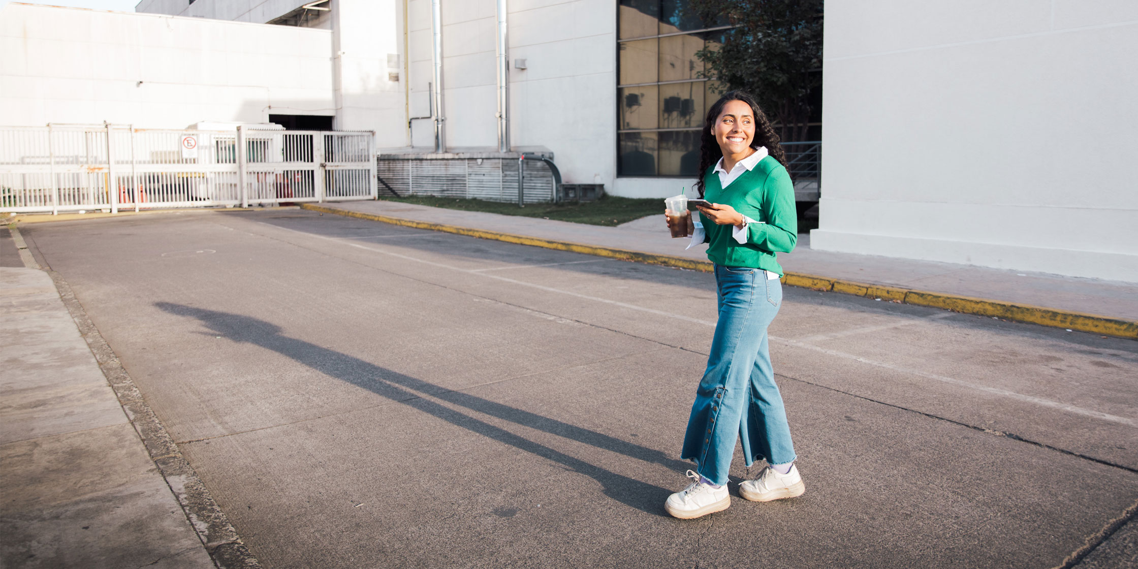 A young woman crosses the street holding coffee and a phone.